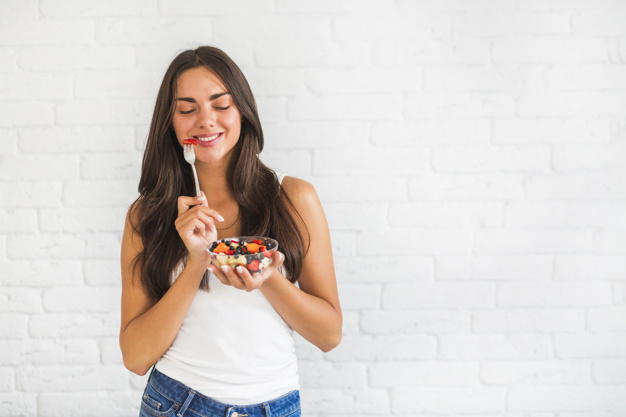 Happy young woman standing against white wall eating fruit salad 23 2147855431